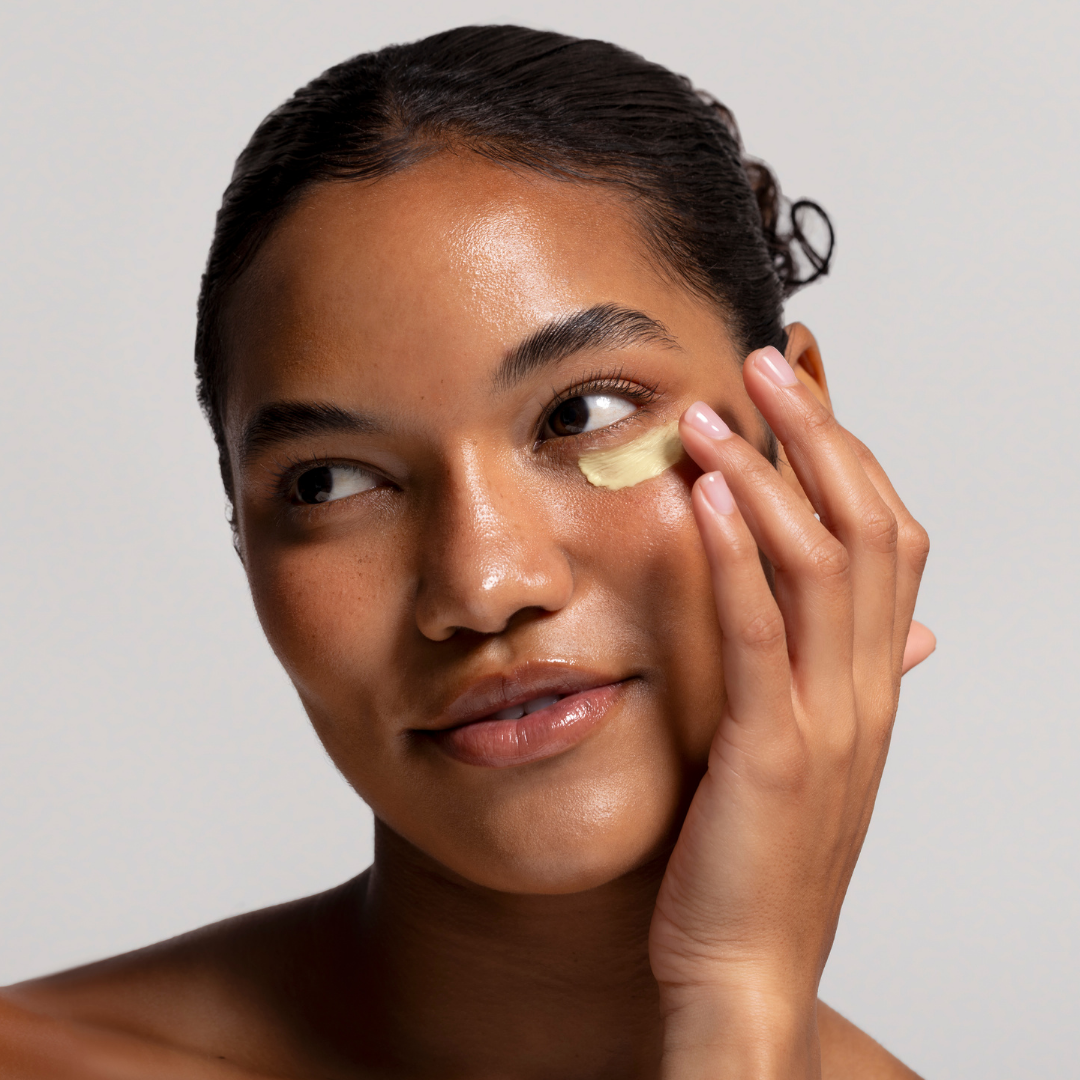Model applying eye cream to her face against a neutral background