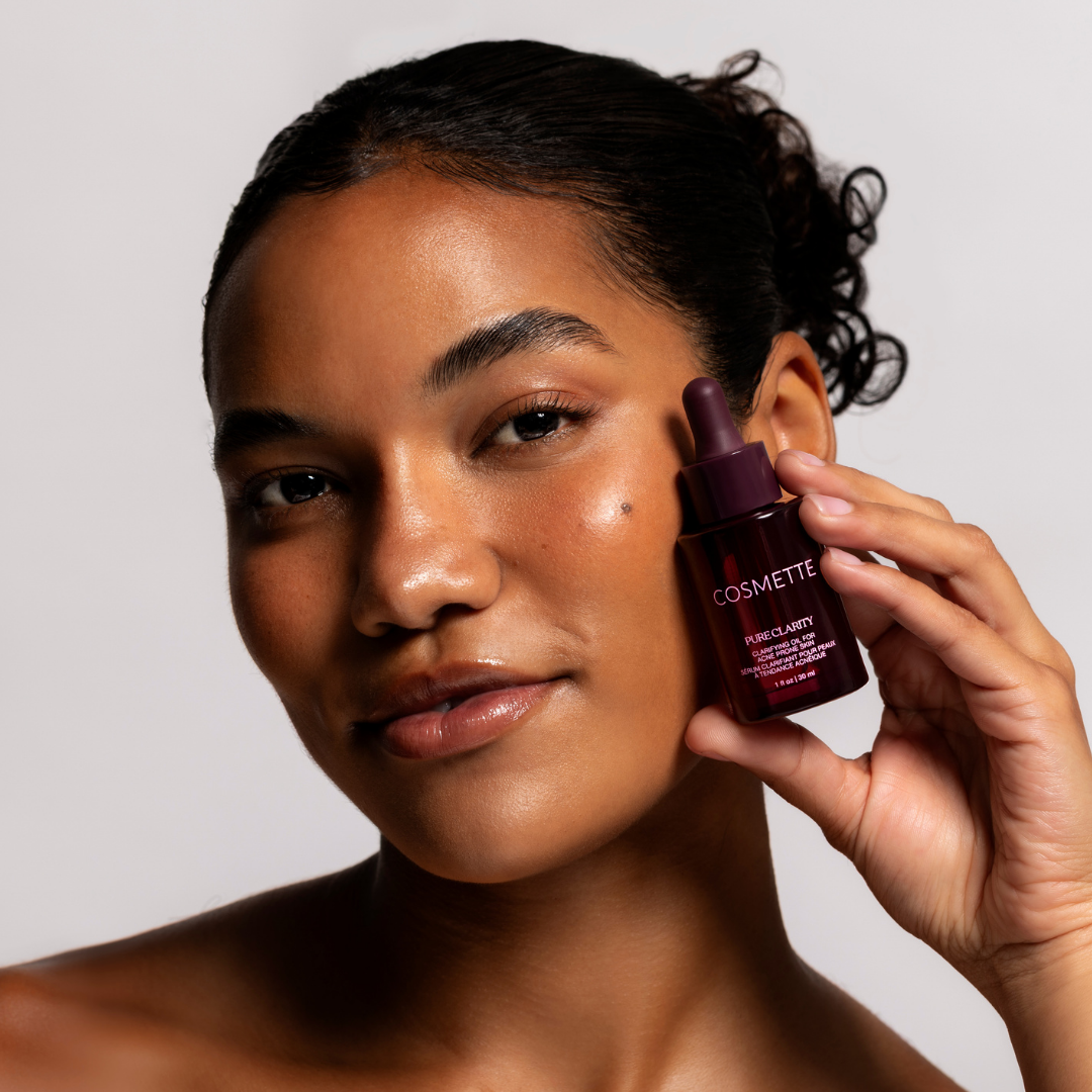 Woman holding a cosmette Pure Clarity Oil dropper bottle near her face against a neutral background