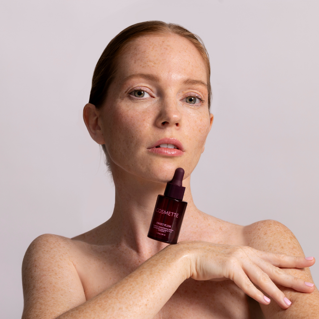Woman holding sweet plum vitamin c serum bottle resting against chin with a plain background